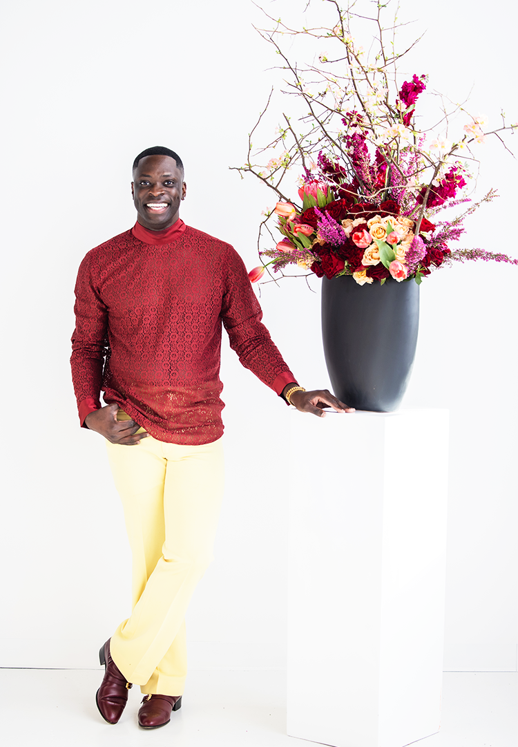 Akeem Clayton of Akeem Clayton Designs posting beside a tall, large floral arrangement contained in Accent Decor's Sasso Vase.