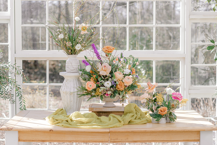 A group image of lush florals designed within a variety of vases and compotes atop a wooden table surrounded by natural light.