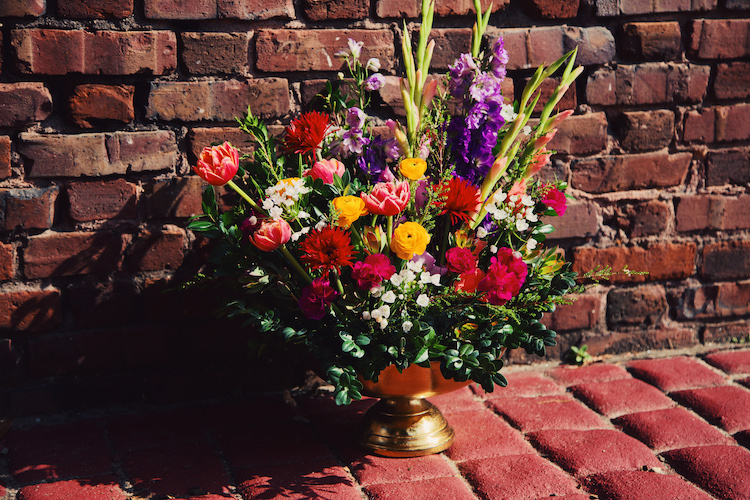 A lush and colorful floral arrangement, styled in Accent Decor's Oscar Bowl, against a brick wall.