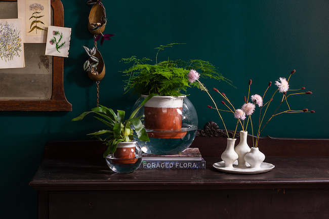 The Davies Watering Planter atop a dard, wooden table surrounded by flowers and home decor accents.