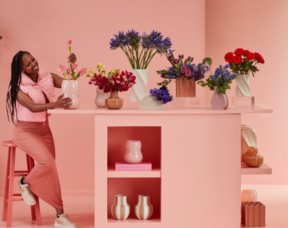A woman sitting in a flower shop surrounded by an array of vibrant blooms. She is nestled among various potted plants and floral arrangements, with a contented smile on her face.