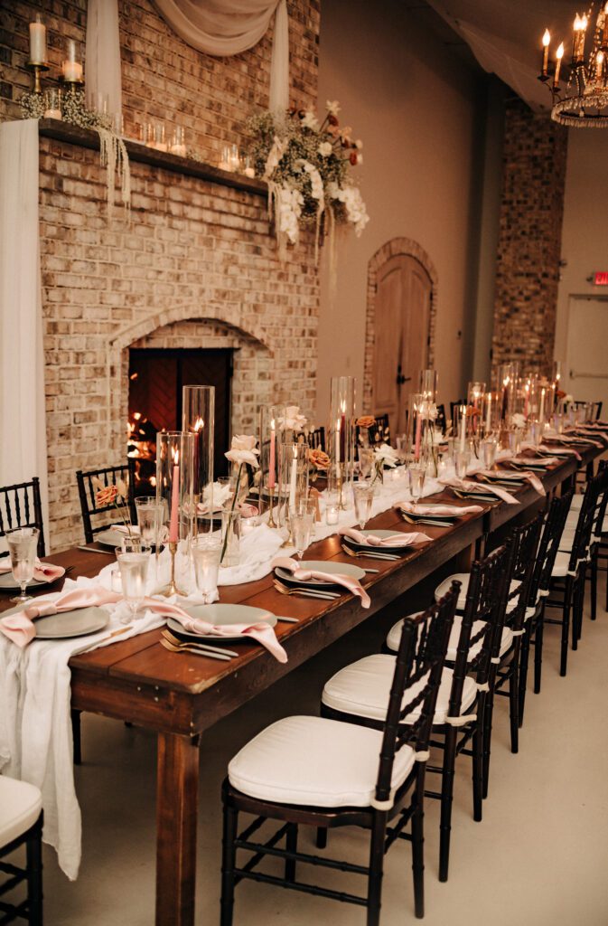 A dramatically decorated long tabletop filled with flatware, lush simple florals and tall glass sleeves.