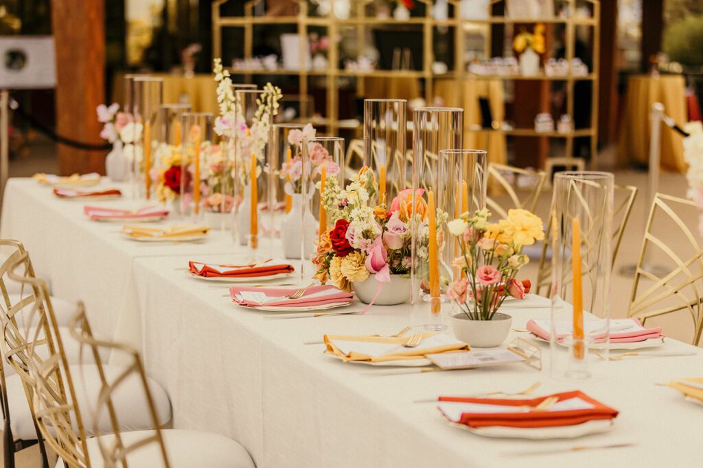 An elegantly decorated long tabletop with colorful flatware, floral centerpieces and tall glass sleeves.