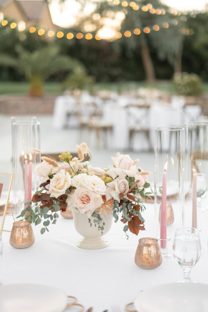 An elegant tabletop decorated with filled with gold and pink hued florals, tall glass sleeves and shimmery votives.