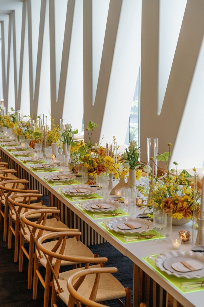 A dramatically decorated long tabletop filled with neon flatware, lush florals and tall glass sleeves.