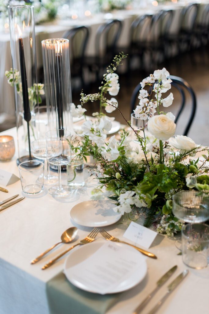 An elegant tabletop decorated with filled with gold and white flatware, lush florals and tall glass sleeves.