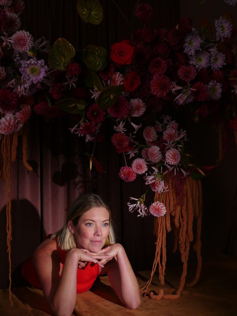 Lys Lytle of Flowering Minds Education poses beneath Accent Decor’s Tenuto Stand, surrounded by lush, cascading florals.