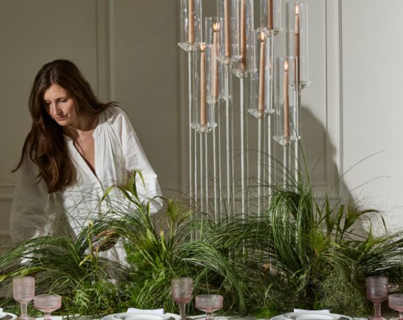 A woman styling an elegant tablescape ft. a grass-inspired centerpiece with wholesale event decor candlesticks, glass drinkware & flatware.