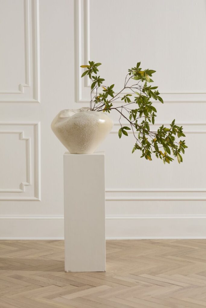 Large white vessel sitting, containing a striking branch with leaves, placed atop a white podium.