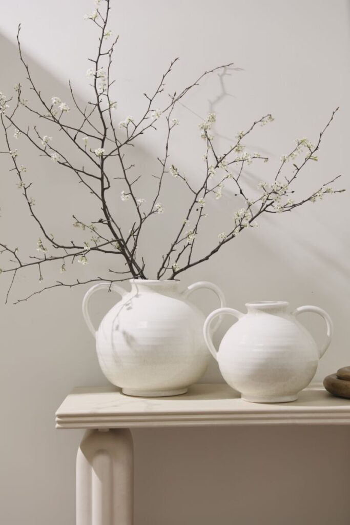 Two large white vessels sitting, one containing a striking branch with white flowers, placed atop a console table.