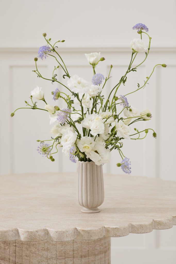 A scalloped wholesale vase atop a console table containing white and lavender florals.