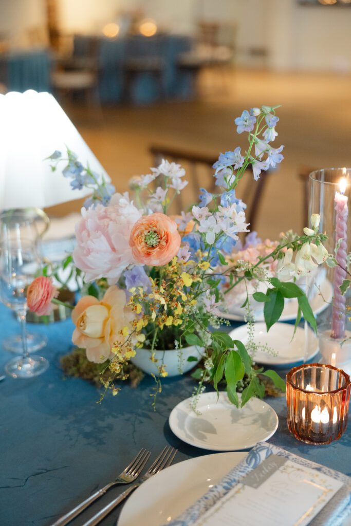 An event tabletop styled with fresh, colorful flowers and wholesale event decor including votives, flatware and a blue tablecloth.