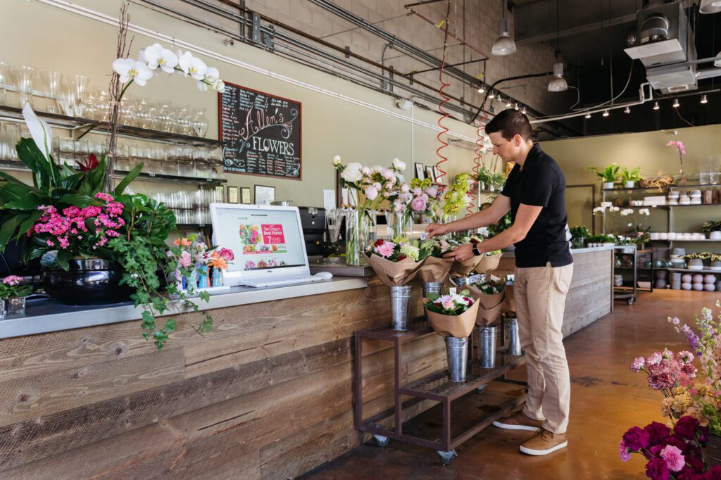 The interior of a floral design studio featuring stocked shelves with vases, fresh floral bouquets and a man at the counter.