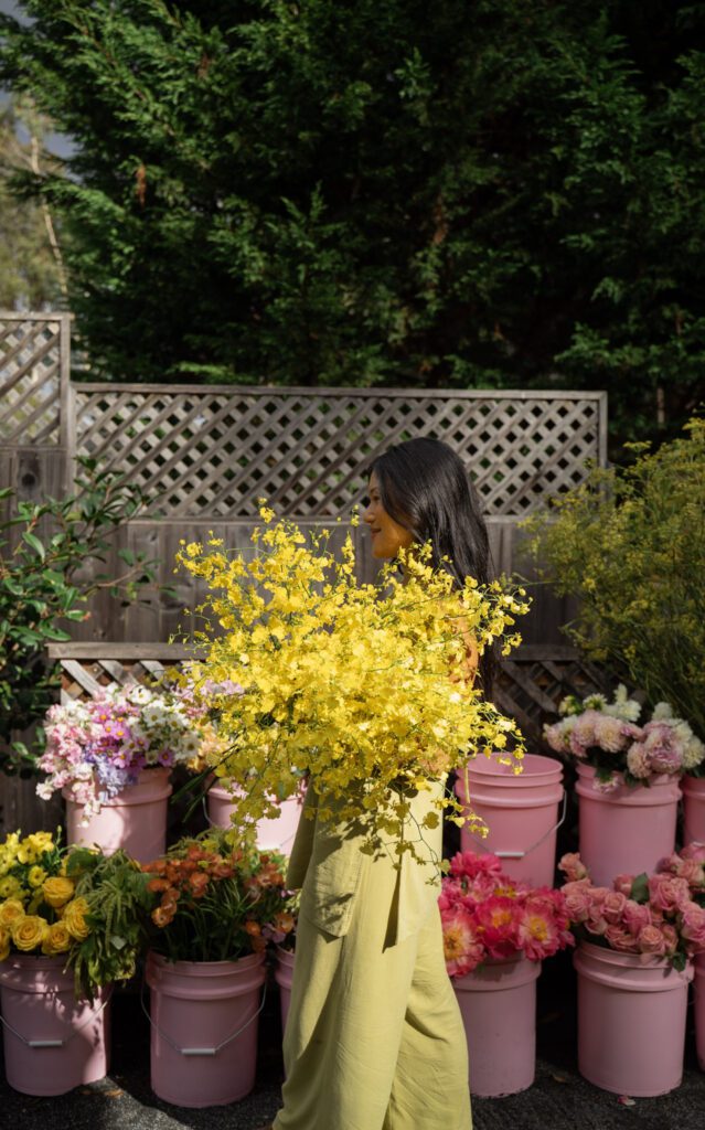 Women holding a lush yellow floral bouquet against a wall of buckets containing colorful flowers.