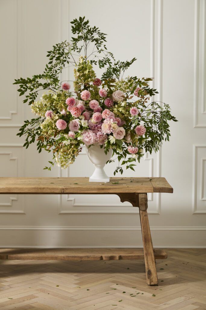 Large Castlebury urn filled with pink-hued florals and lush greenery, styled atop a rustic wooden bench.